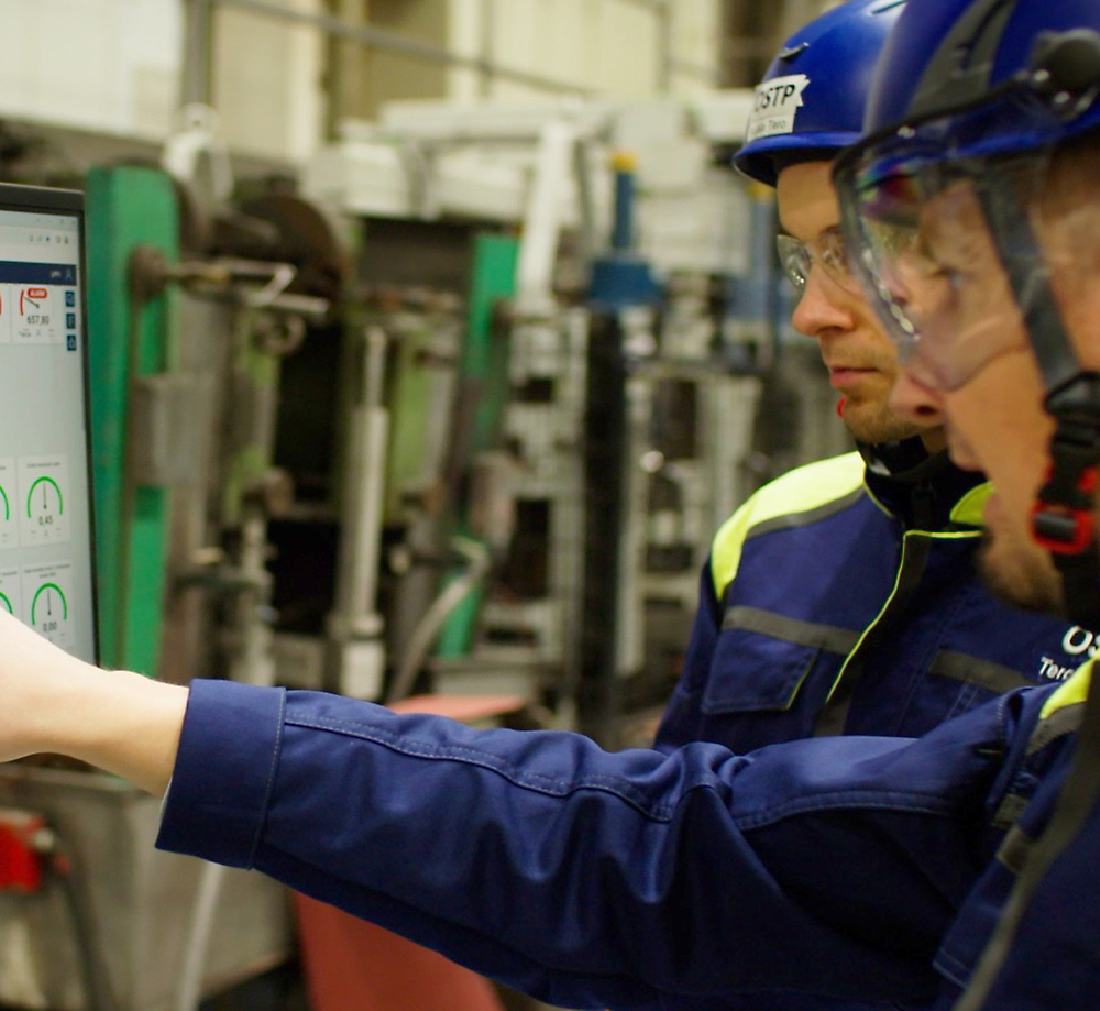 Two factory workers wearing safety gear are discussing information displayed on a screen in an industrial setting.