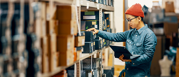 A man in a denim jacket and red beanie reviews inventory with a tablet in a warehouse aisle.