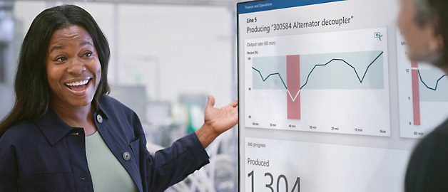 A smiling woman presenting a production graph on a digital screen to a colleague in an office setting.