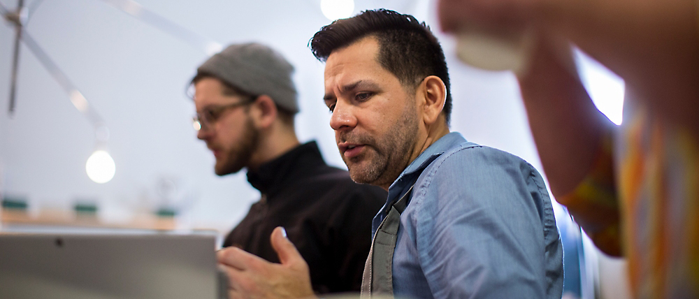 Two men in an office setting, one focused on a laptop and the other engaging in a conversation