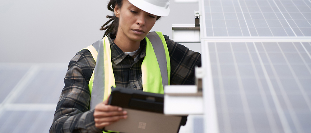 Una mujer técnica con un casco y un chaleco de seguridad inspecciona un panel solar con un dispositivo digital.