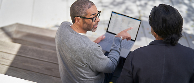 Two men examining a laptop screen together outdoors, one pointing at the screen while engaging in a discussion.