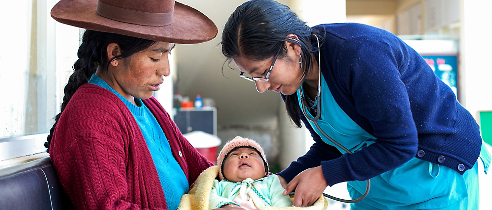 עובד בריאות בוחן תינוק שמחזיקה אישה בלבוש מסורתי בתוך מרפאה.