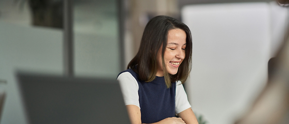 En smilende asiatisk kvinne i blå og hvit topp som sitter ved et skrivebord på et kontor, og samhandler med en kollega vi ikke kan se.