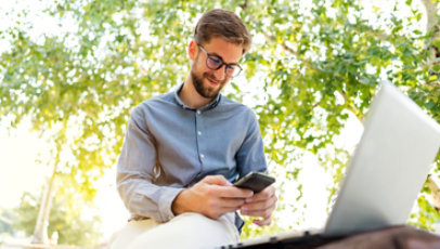 A man sitting outside happily checking his digital phone and laptop with the sun shining over his shoulder.