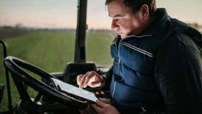 A man seated outdoors behind the wheel of a farming vehicle looks at a tablet device.