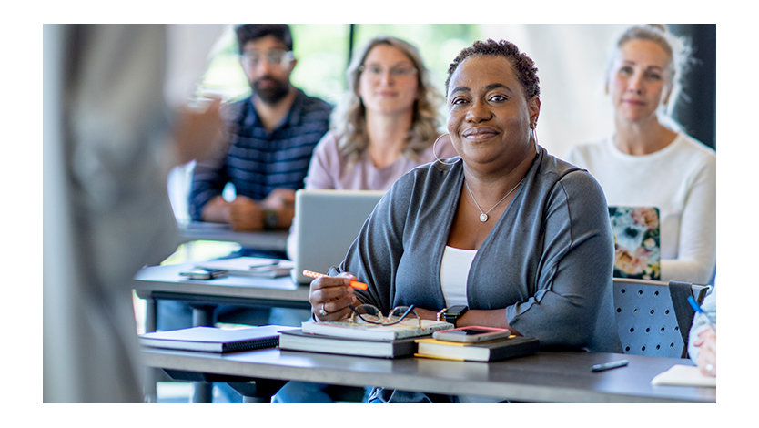 Adult woman attentively listening to a instructor in a classroom surrounded by adult students on laptops.