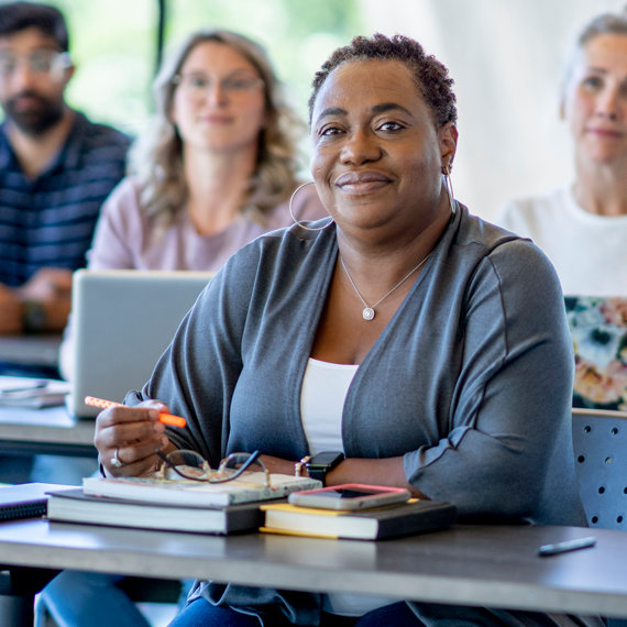 Adult woman attentively listening in a classroom surrounded by adult students on laptops.