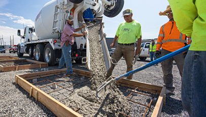 Construction workers pour concrete from a large mixing truck into a frame on the ground.