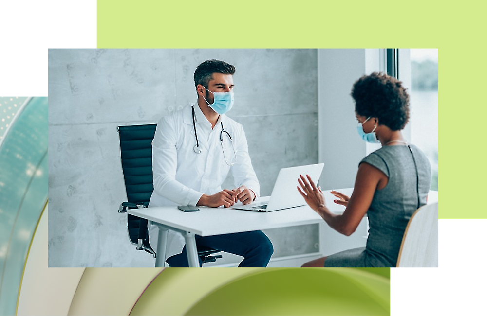 A doctor wearing a mask and sitting at a desk with a laptop and talking with another person.