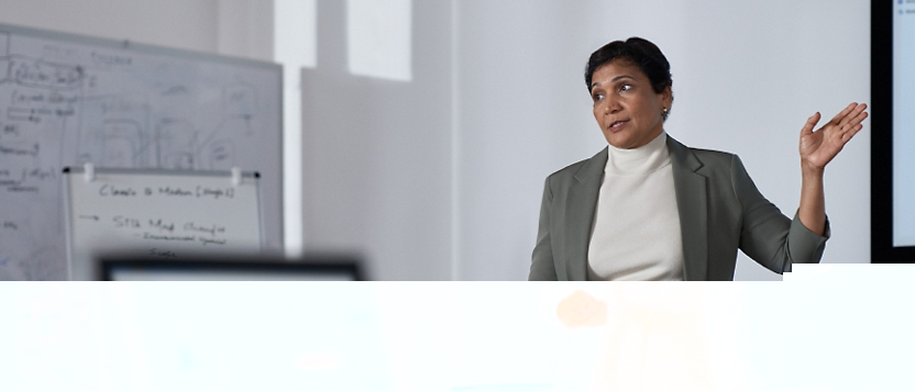 A professional woman presenting in a meeting room with a whiteboard and a computer in the background.