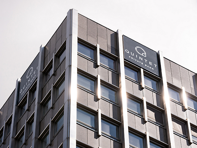 Moderno edificio de oficinas gris con el logotipo del "Quintet" en la parte superior, bajo un cielo despejado, la luz del sol reflejándose en las ventanas.