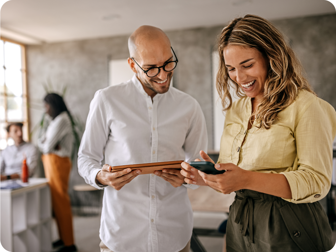 Two colleagues, a bald man and a woman with long blonde hair, smile while looking at a smartphone together