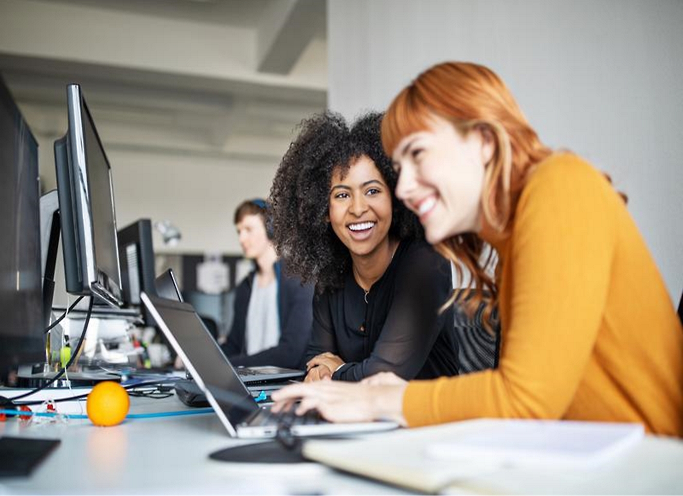 Zwei lächelnde Frauen, die am Schreibtisch sitzen und auf den Laptop blicken.