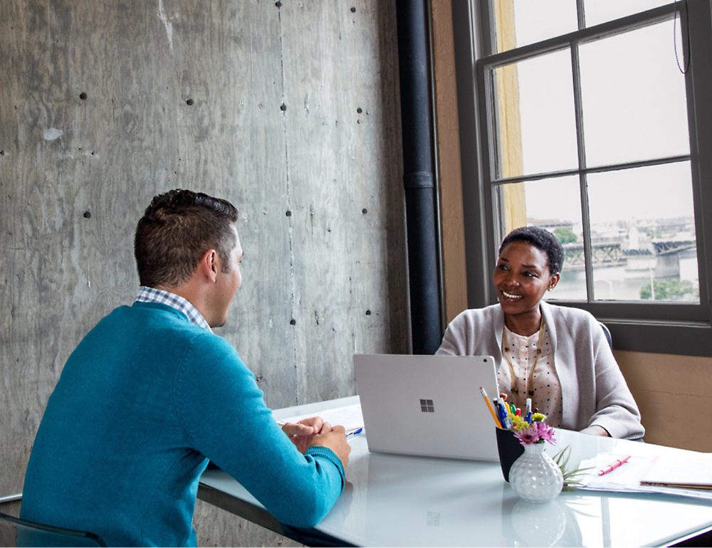 Two professionals sitting at a table in a modern office, conversing over a laptop. the woman is smiling.