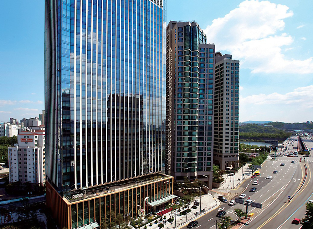 Modern cityscape showing a tall glass office building next to residential towers over a busy street under a clear blue sky.