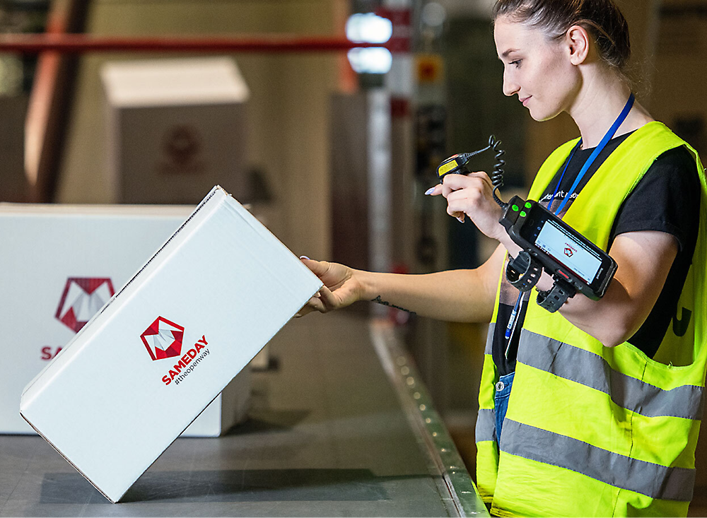 A female worker in a high visibility vest scans a barcode on a package in a warehouse.