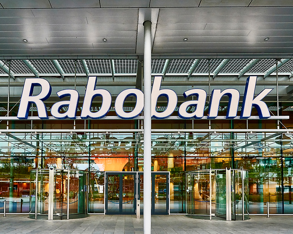 Vista exterior de un rabobank con puertas de cristal y una gran señal que muestra el nombre del banco encima de la entrada.