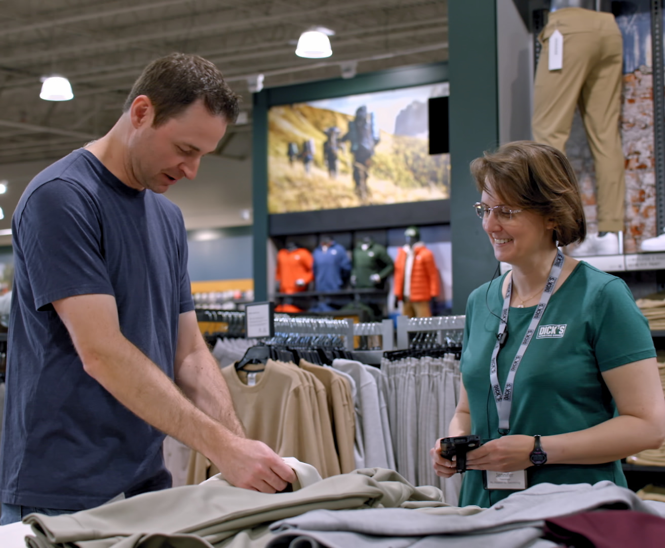 En man som undersöker kläder vid en butiksdisk med hjälp av en leende kvinnlig anställd som bär en namnskylt.