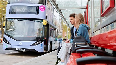 Två personer sitter på en bänk bredvid en tvåvåningsbuss.