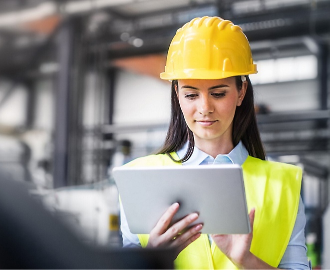 En kvinde iført hjelm bruger en tablet på en fabrik.