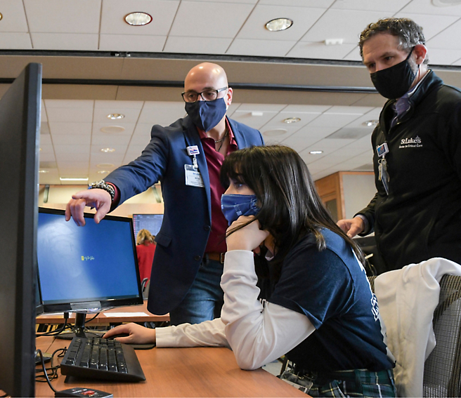 A person is pointing at a computer screen while another person, wearing a mask, observes