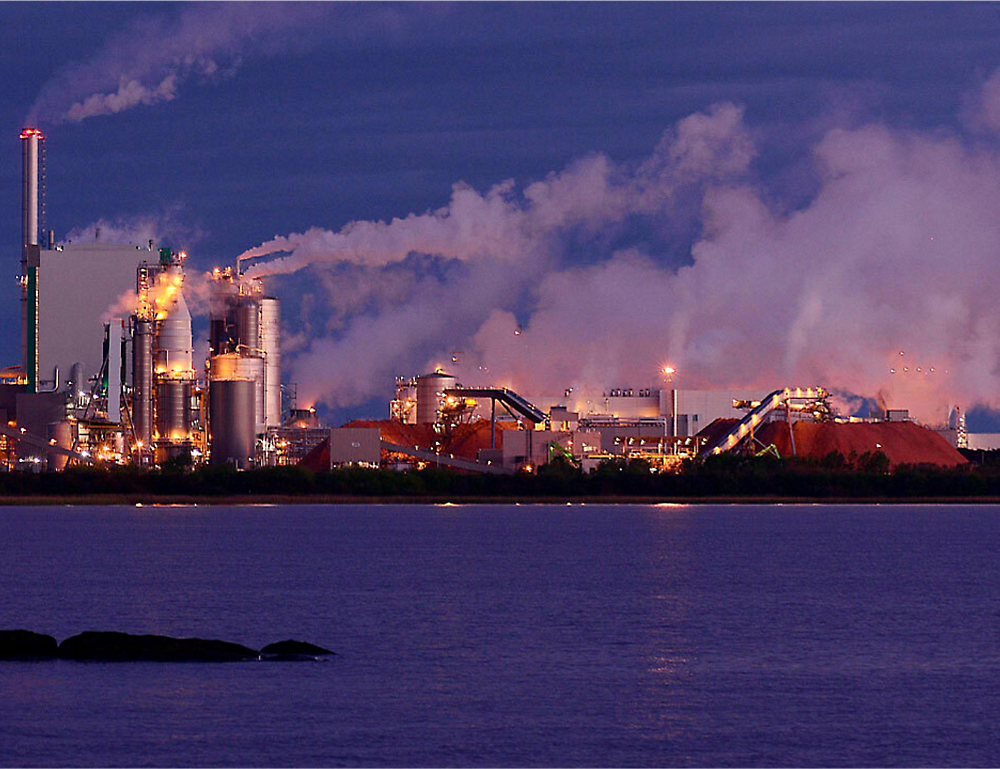 Industrial facility with multiple smokestacks emitting smoke, situated next to a body of water.