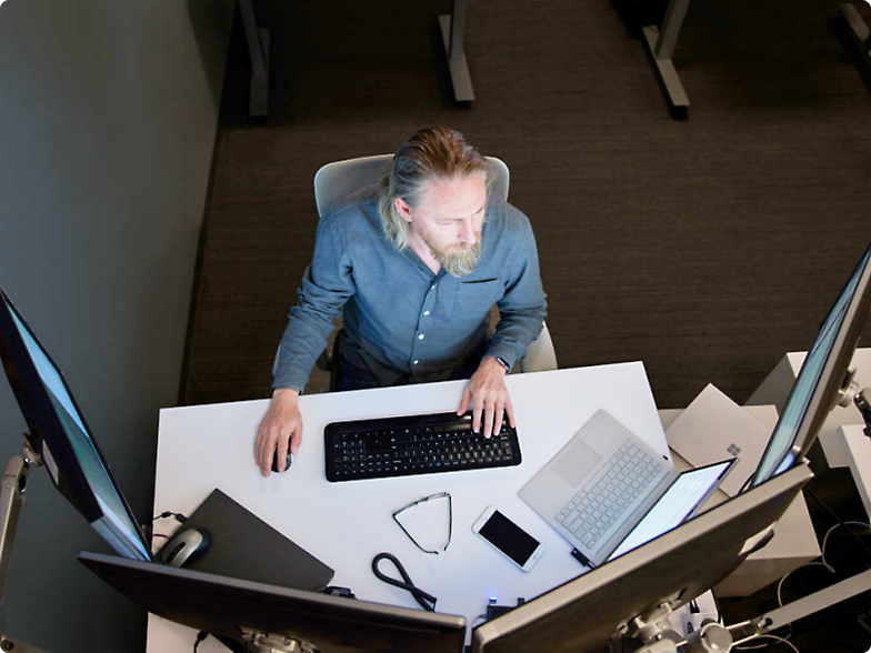 A man sitting at an office workstation, using a keyboard with multiple monitors and a laptop on the desk.