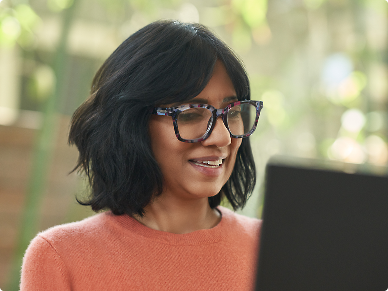 A smiling woman with glasses, wearing a peach sweater, looks at her laptop screen in a sunlit room.