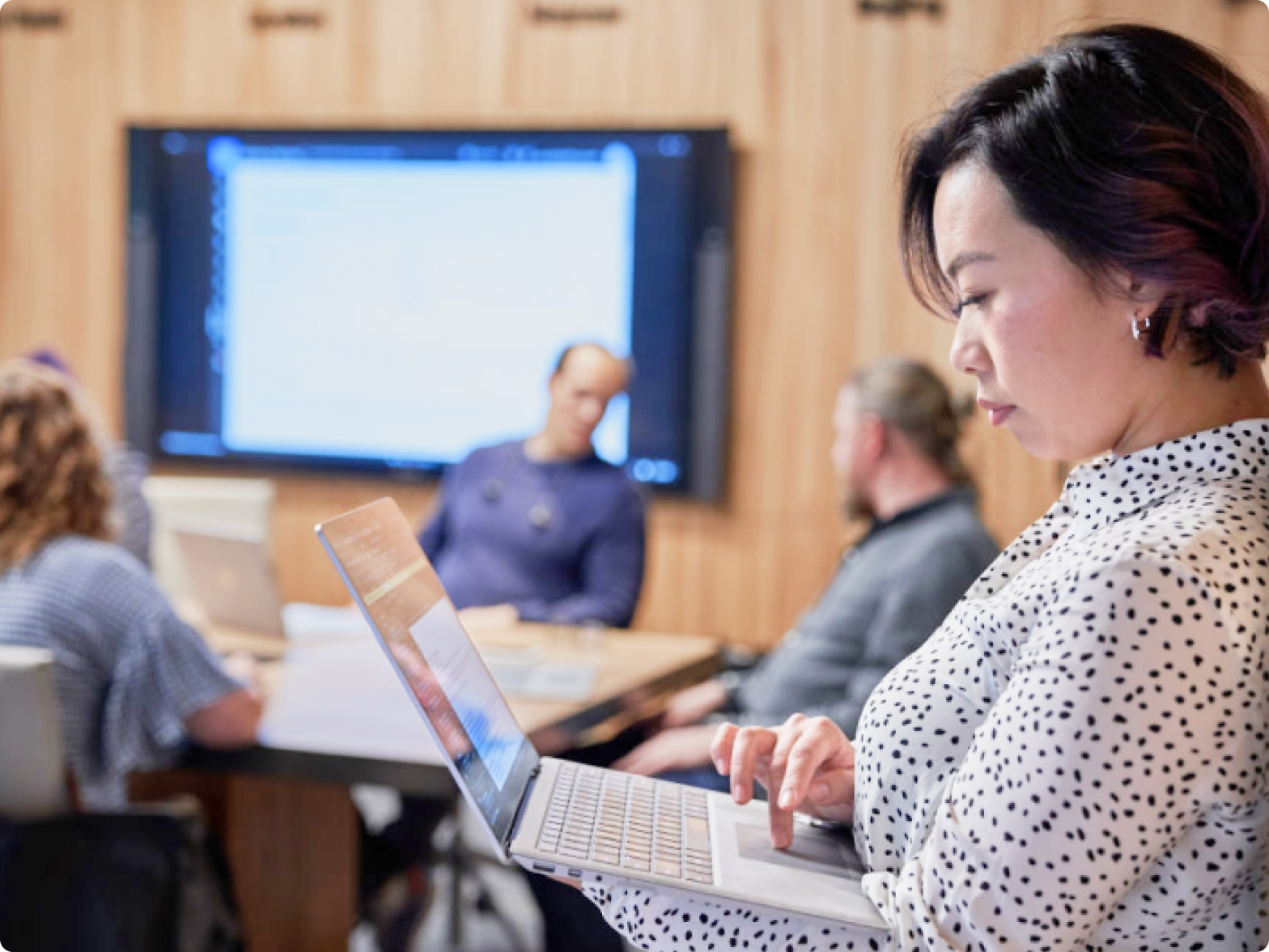 A woman using a laptop in a conference room with three other colleagues and a presentation screen in the background.