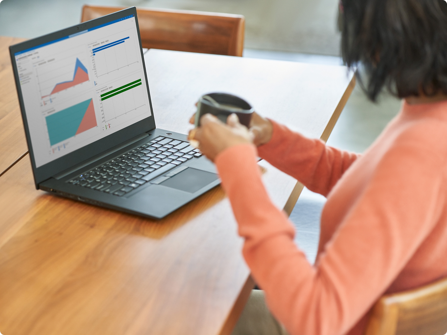 Frau in einem orangefarbenen Pullover an einem Holztisch, die mit einem Laptop Diagramme anzeigt und eine Kaffeetasse hält.