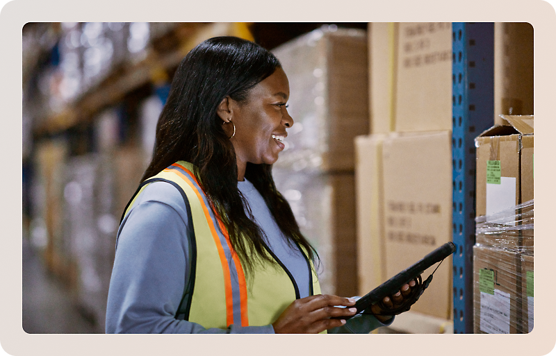 A warehouse worker wearing a safety vest uses a tablet while standing next to shelves filled with boxes.