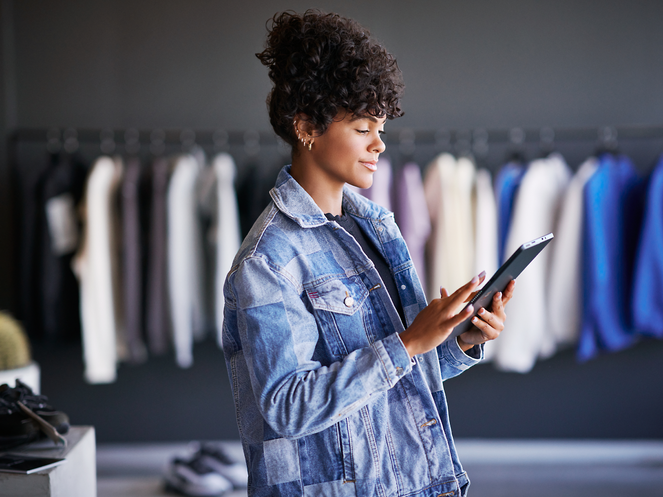 A person wearing a denim jacket is looking at a tablet in a clothing store, standing in front of a rack of hanging garments.