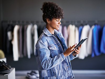 A woman in a denim jacket is using a tablet in a clothing store.