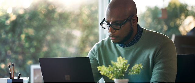 Eine Person mit Brille und grünem Pullover arbeitet an einem Laptop an einem Schreibtisch