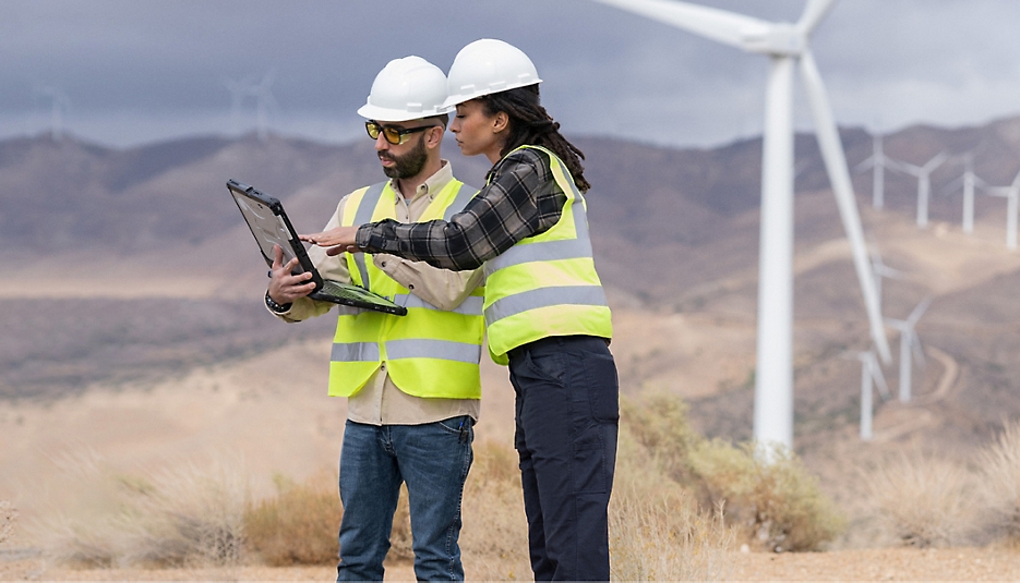 Twee mensen met veiligheidshelmen en veiligheidsvesten staan in een veld met windturbines en kijken naar een laptop