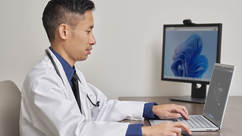 Медицински специалист, който носи бяла престилка и стетоскоп и седи на бюро с лаптоп, свързан с настолен монитор