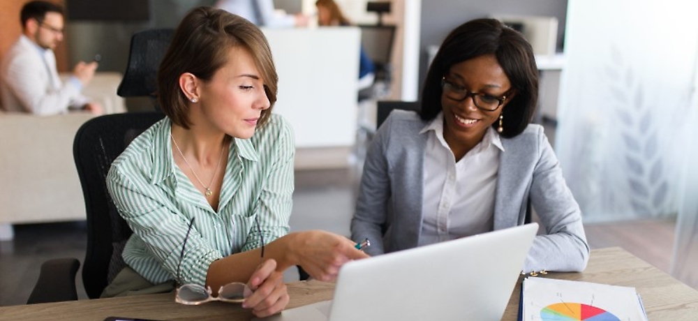 Two women, one caucasian and one african american, collaborating over a laptop in a modern office setting.