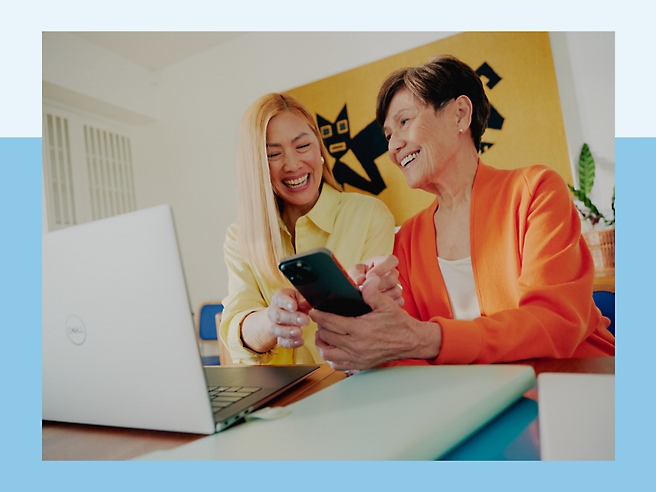 Two women smiling while looking in the phone while a laptop is open on the table