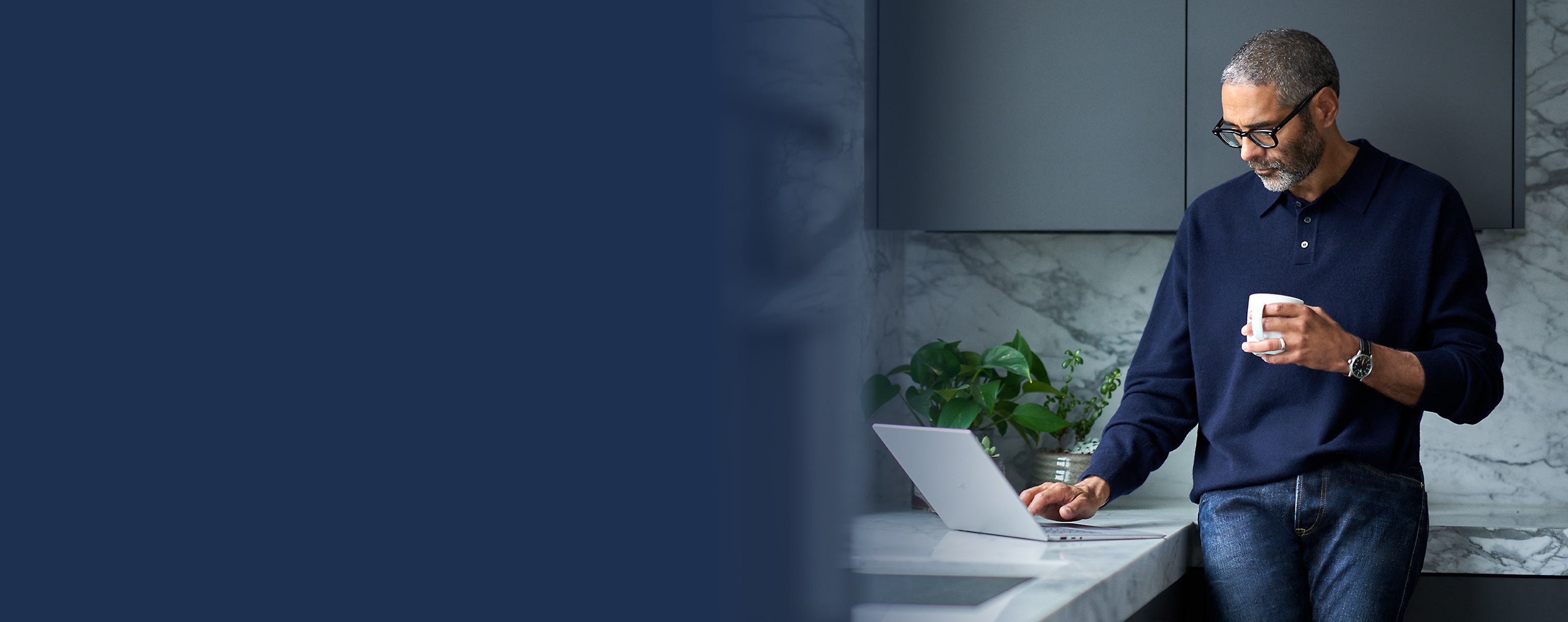 A man with glasses holding a mug while using a laptop in a kitchen setting.