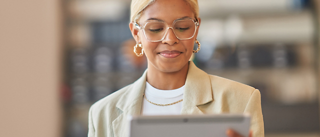 A woman with glasses and earrings, wearing a light jacket, reads content on a tablet with a focused expression.