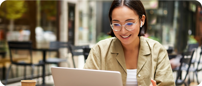 A person with glasses and earphones sits at an outdoor cafe, smiling at a laptop screen.