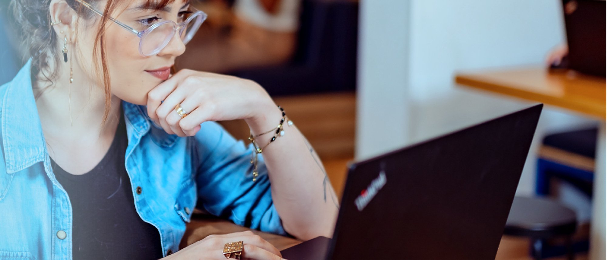 O femeie cu ochelari, lucrând pe laptop cu o mână pe bărbie