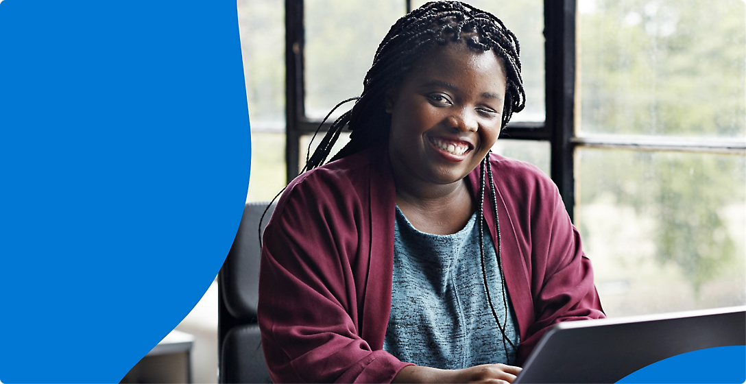 A smiling black woman with braided hair, wearing a burgundy cardigan, sits at a desk near a window, using a laptop.