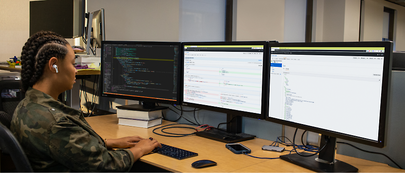 A person in military attire working on multiple computer monitors displaying code and data.