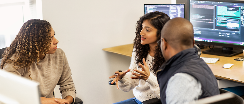 Three professionals discussing a project near a computer monitor in an office setting.