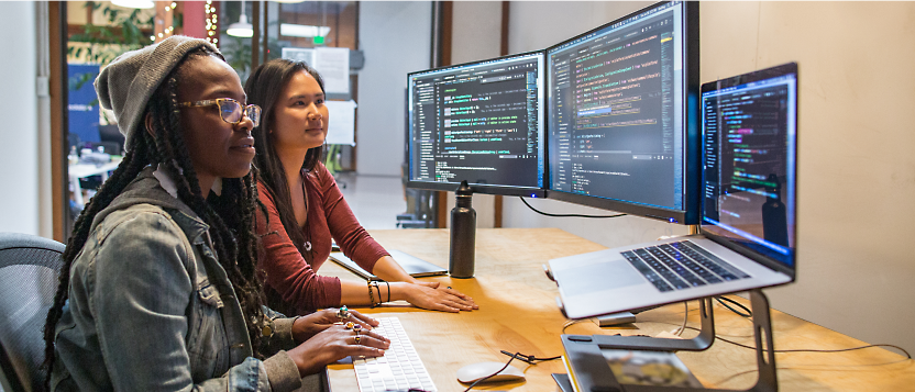 Two individuals collaboratively working at a computer workstation with multiple monitors displaying code.