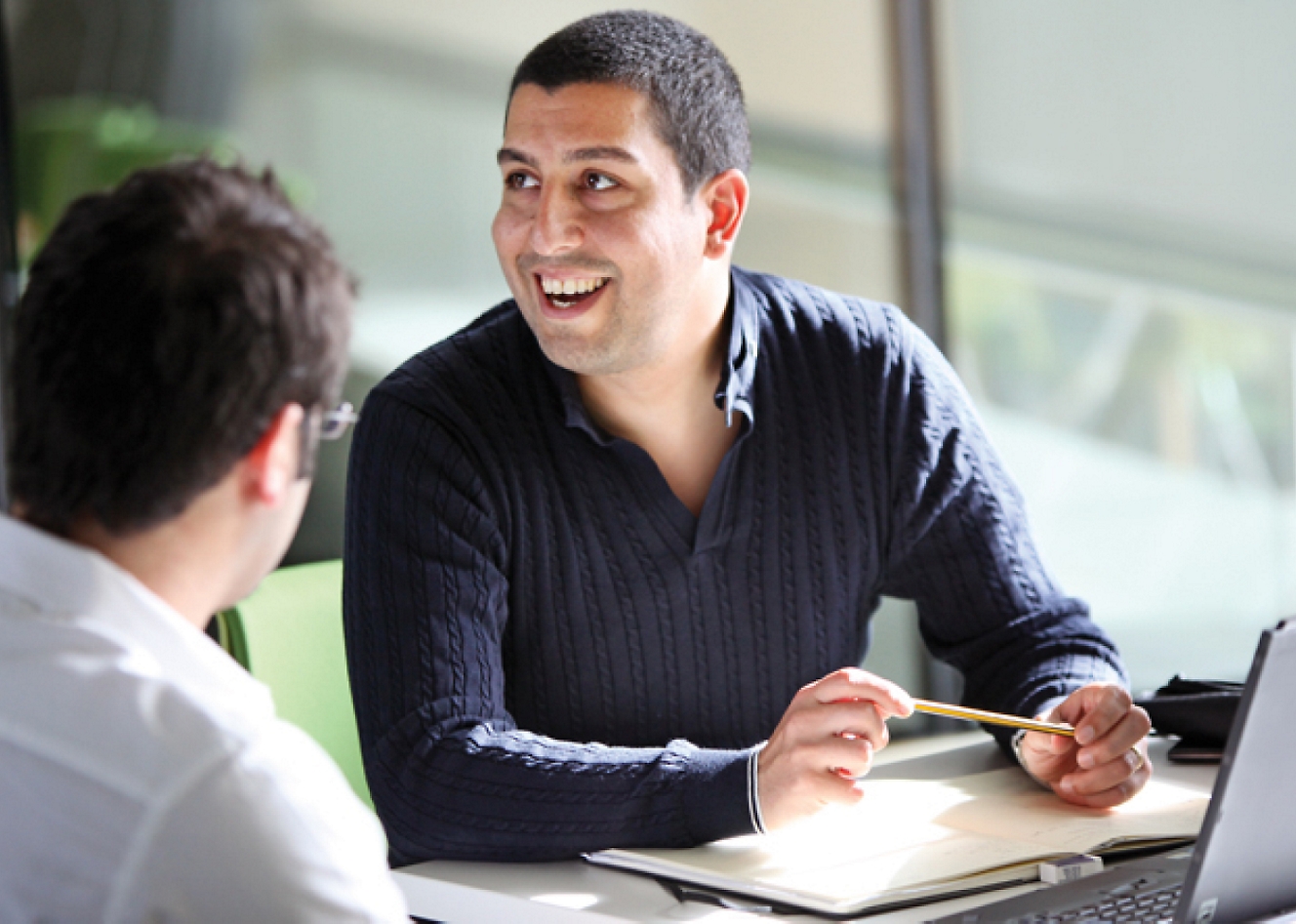 Two men sitting at a desk talking to each other.