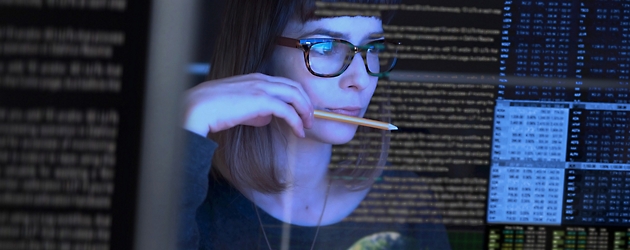 Femme avec des lunettes examinant du code sur plusieurs écrans d’ordinateur dans une pièce sombre, tenant un crayon à sa bouche.