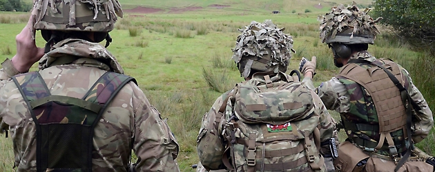 Drei Soldaten in Tarnkleidung stehen auf einem Feld, von der Kamera abgewandt, und beobachten die Landschaft.